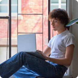 Một thanh niên ngồi bên cửa sổ đang mở, tay cầm máy tính
