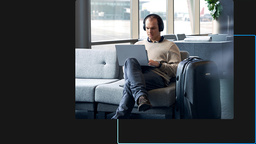 A person sitting in an airport lounge wearing headphones and using their laptop with their legs crossed and their suitcase nearby