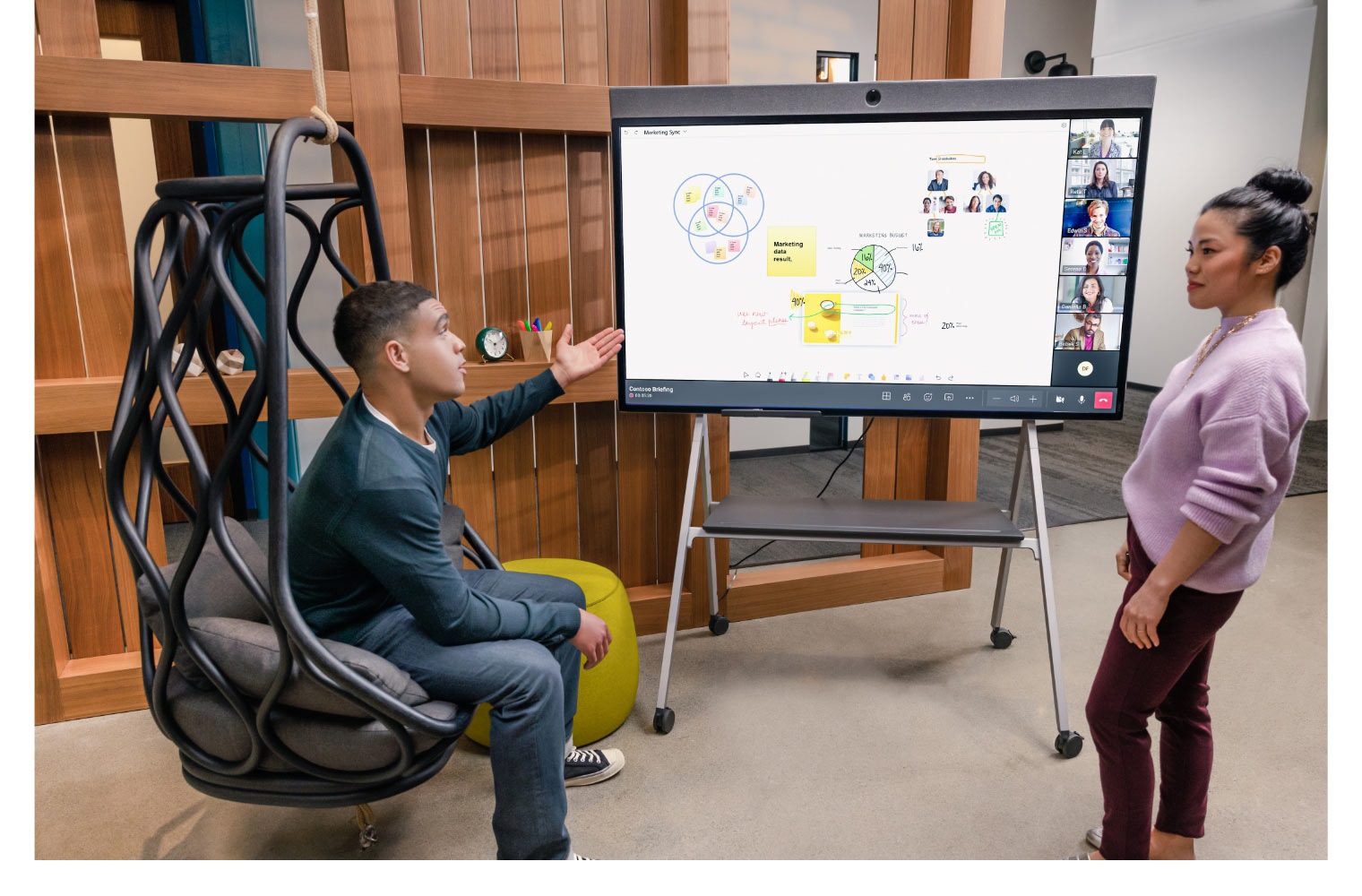 A person sitting in an egg-shaped chair having a conversation with a coworker while participating in a Teams meeting being displayed on a large screen next to them where a collaborative whiteboard is being edited in real time.