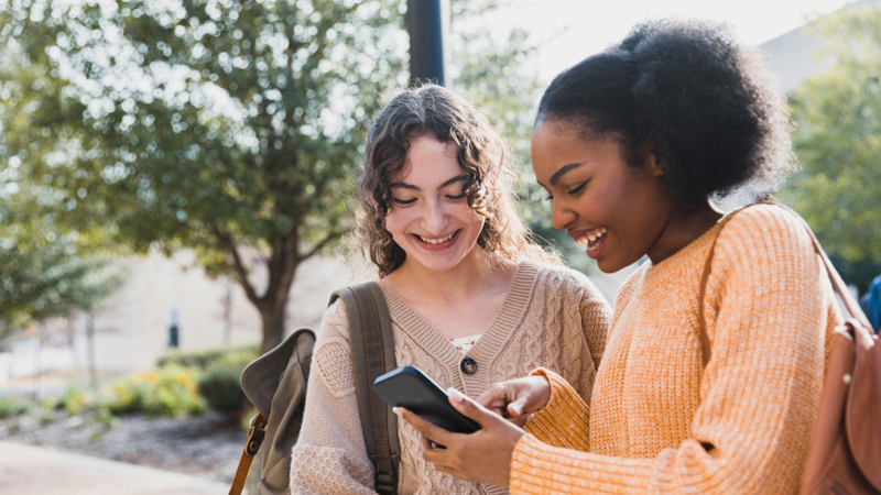 Two teenage girls standing outside looking at a smartphone and smiling.