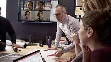 One person speaks at a conference room table as four people look on via a screen on Microsoft Teams and several others watch from in the room.
