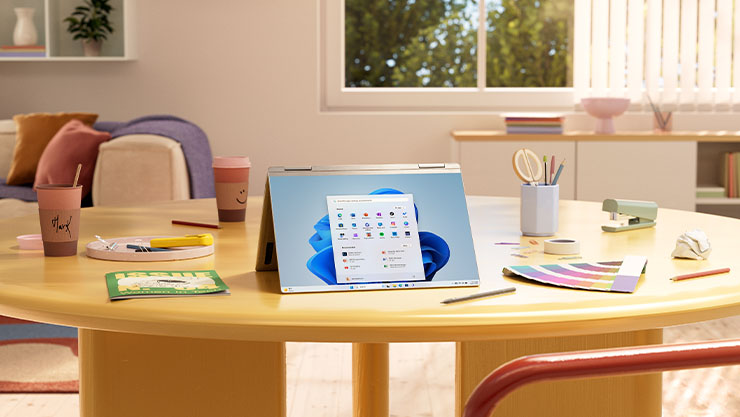 A folded PC with a Windows 11 bloom screen and start menu stands on a wooden table with various office supplies surrounding