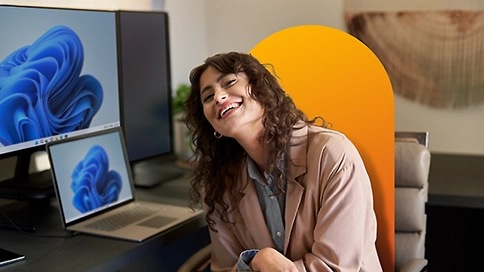 Woman smiling sitting at a desk with Windows 11 on laptop and desktop