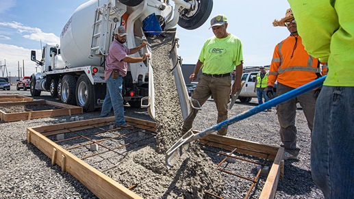 Construction workers pour concrete from a large mixing truck into a frame on the ground.