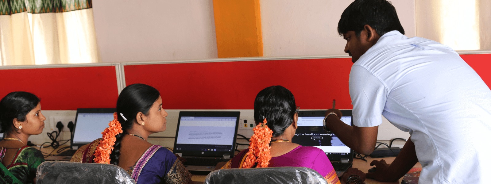 Dans un laboratoire informatique, un enseignant s’adresse à trois étudiants qui se tiennent face à leur ordinateur portable.