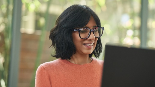 A woman wearing glasses works on a laptop in an office environment.