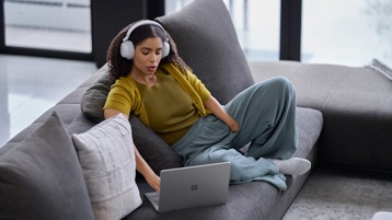 A person with headphones on uses laptop while sitting on a couch.