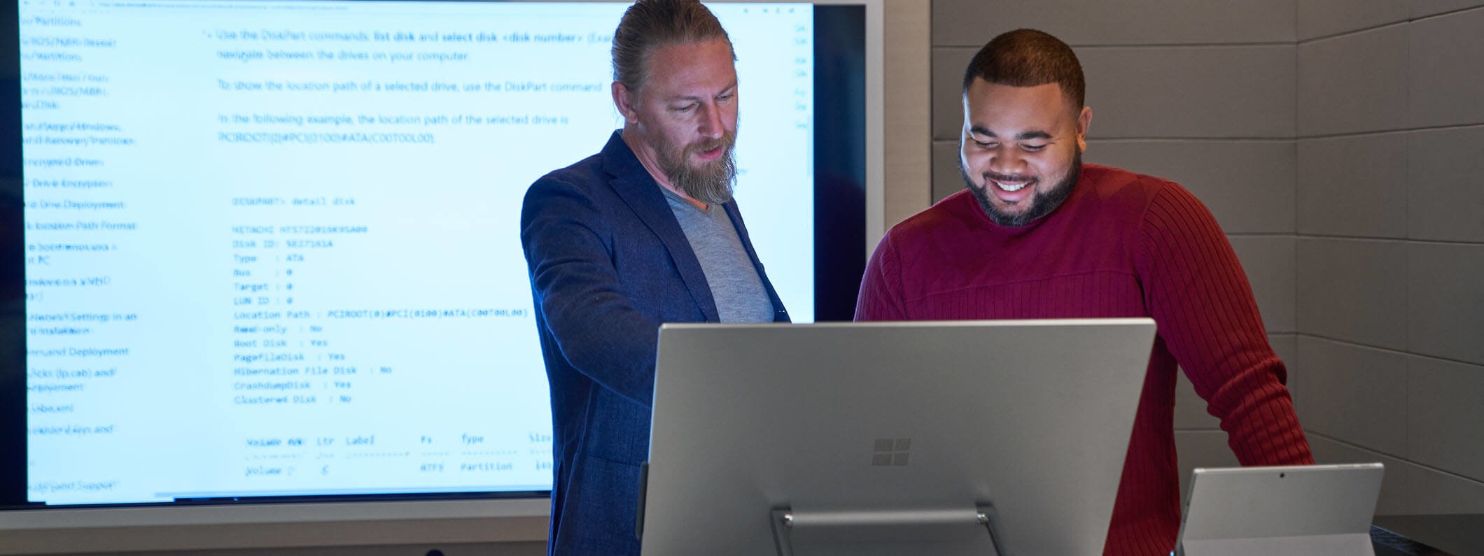 Forward facing view of two men working on a Microsoft Surface Studio with a larger blurred screen/display behind them.