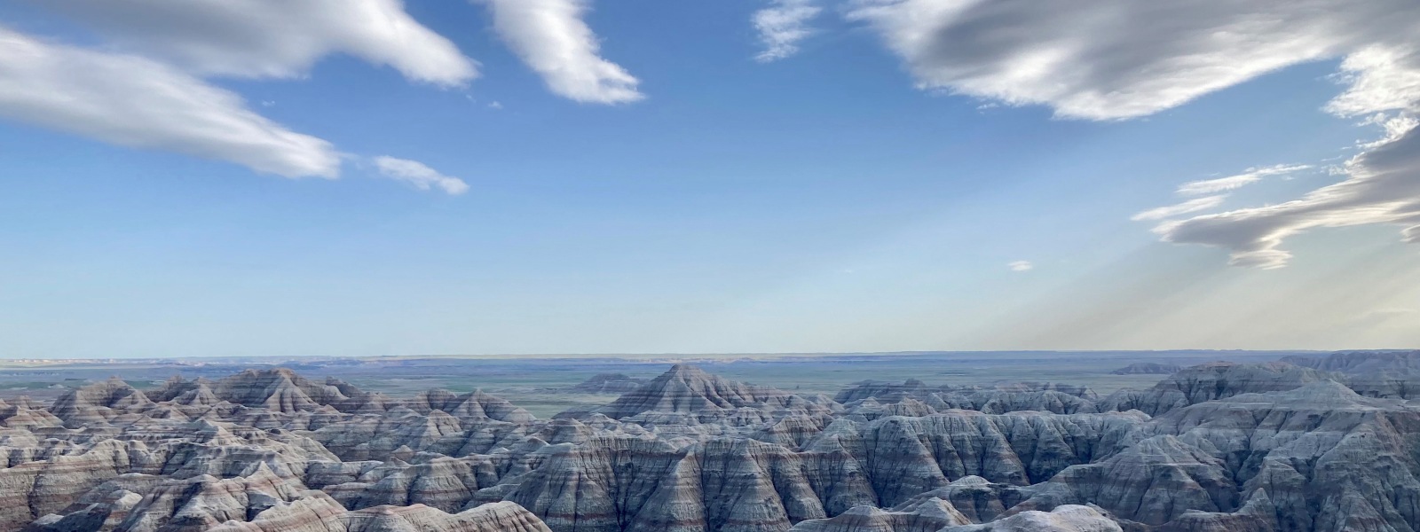 Landscape of mountains in the Badlands National Park with distinguished bands of color throughout the formations, blue skies with some clouds present.