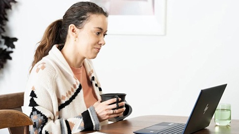 Persona sosteniendo una taza de café y mirando un equipo portátil que tiene en frente.