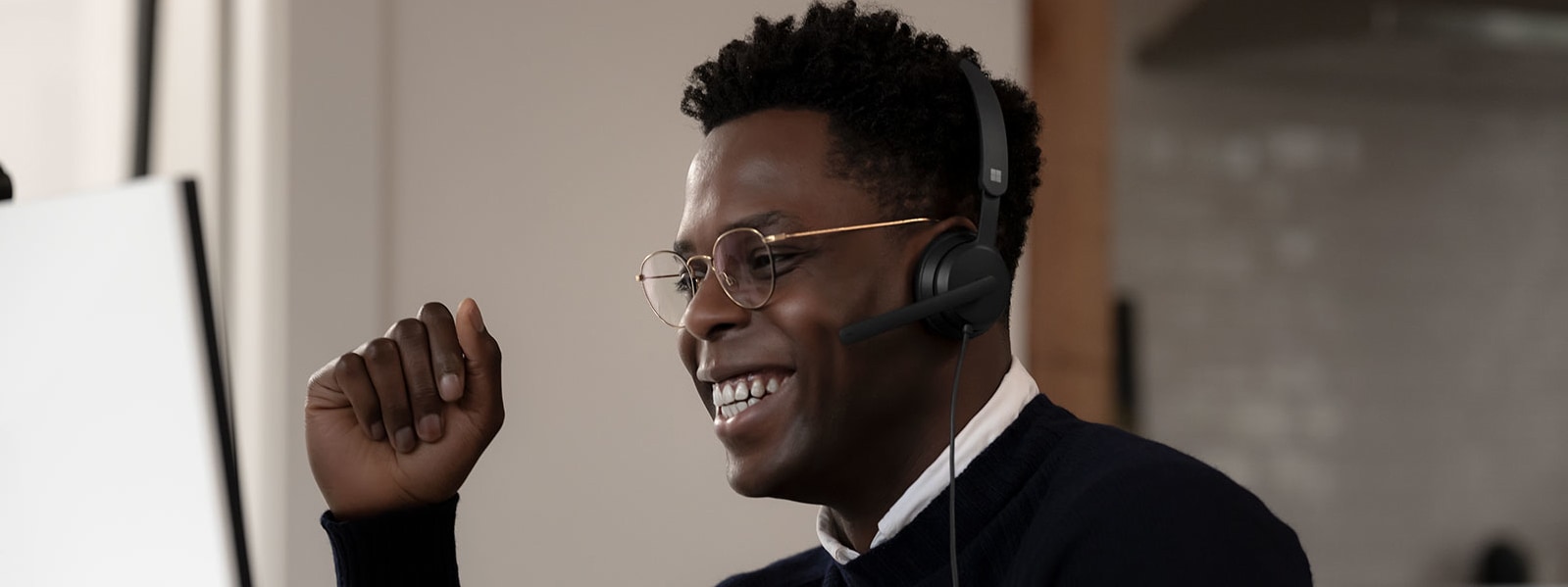 Un hombre lleva puestos unos audífonos Microsoft Modern USB headset, sonríe hacia su equipo mientras levanta la mano