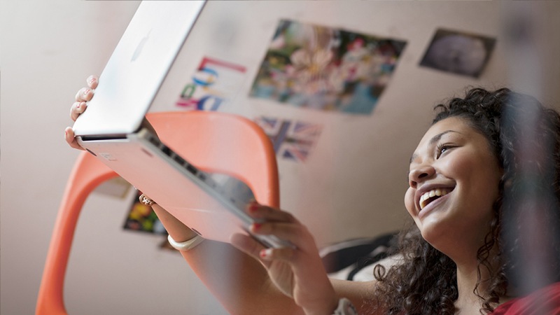 A teen smiles and holds up a laptop in her bedroom.