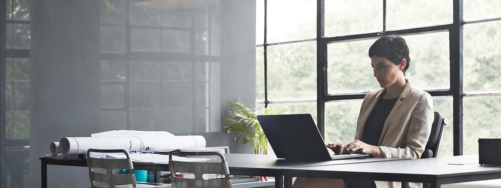 Uma mulher está sentada a uma mesa longa e digitando em um laptop em um escritório moderno com paredes de vidro.
