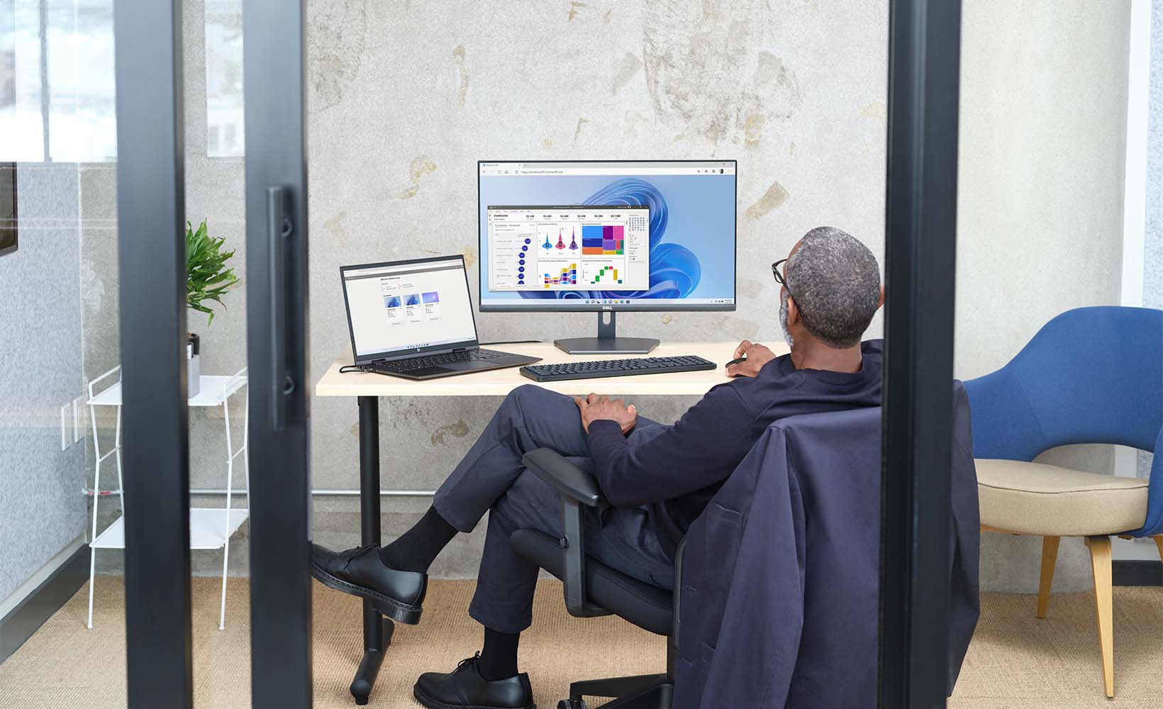 A person sitting in a glass office using a laptop connected to a desktop monitor.