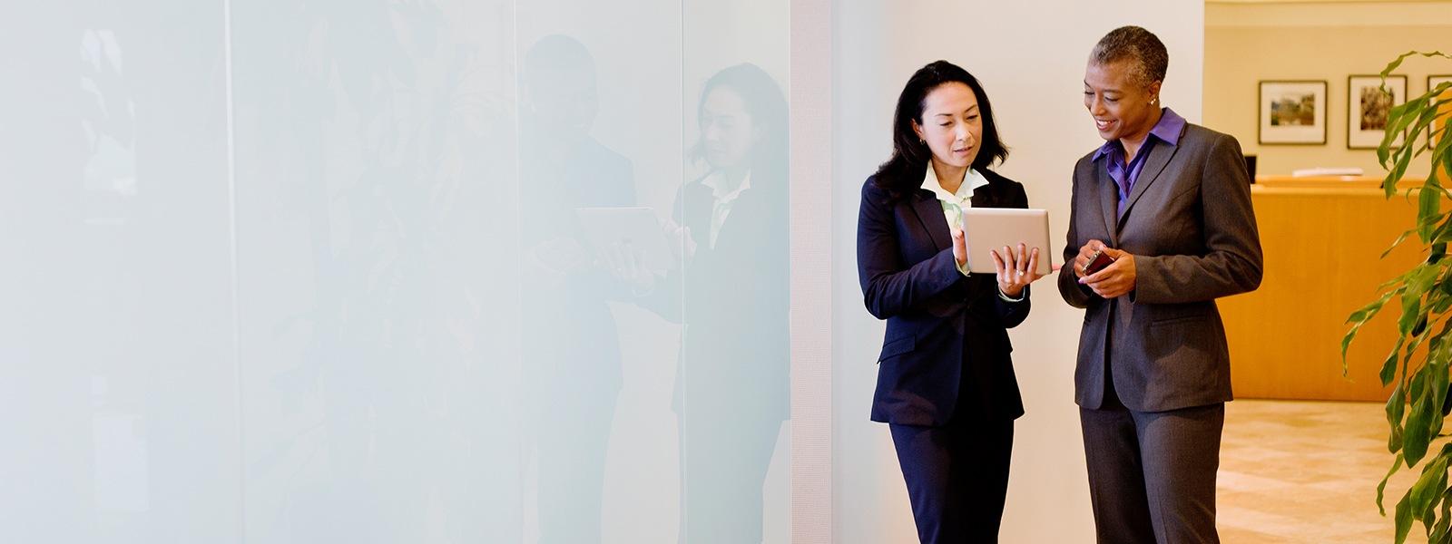 Two businesswomen dressed in suits are using a digital tablet while chatting in an office corridor.