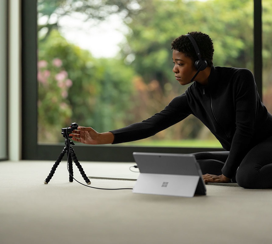 A female wearing a Microsoft Modern Wireless headset leans over to adjust a Microsoft Modern webcam that’s next to a Surface Pro laptop