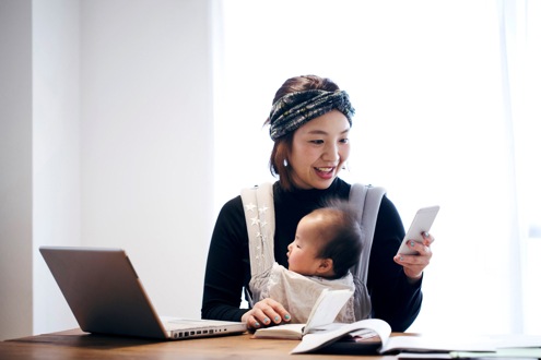 Japanese woman in casual clothes carrying her baby in baby carrier and using a smartphone on the desk. She‘s working and doing childcare at home.