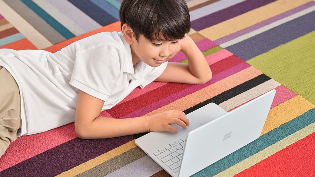 カーペットでくつろぎながら Surface Laptop SE を使用する子ども。