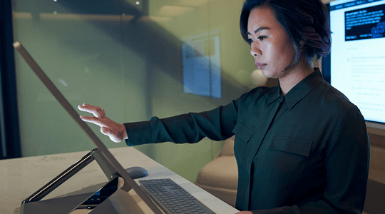 Perfil lateral de uma mulher a usar uma camisa escura num escritório escuro a rolar ou a trabalhar num Microsoft Surface Studio.