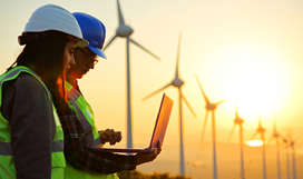 Two workers wearing safety vests and hard hats looking at a laptop and standing in front of large wind turbines.