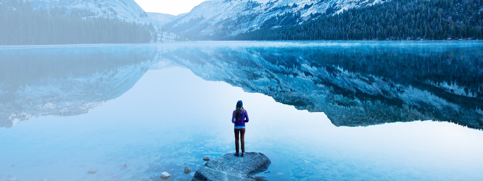Woman stands on the shore of a lake surrounded by snowy mountains