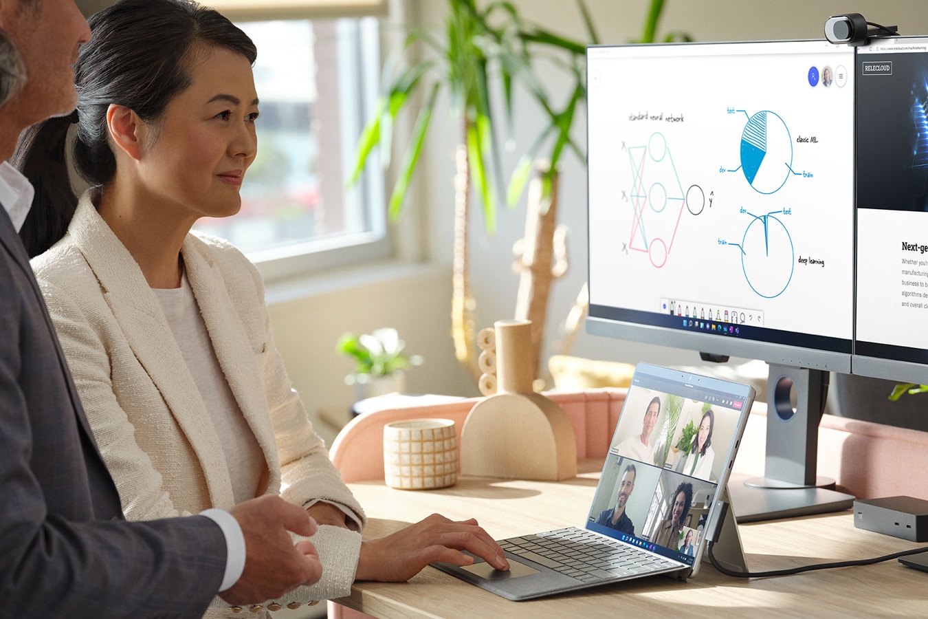 Un bărbat și o femeie într-un birou folosesc dispozitivul Surface, conectat la două monitoare externe, pentru a conduce o ședință Microsoft Teams
