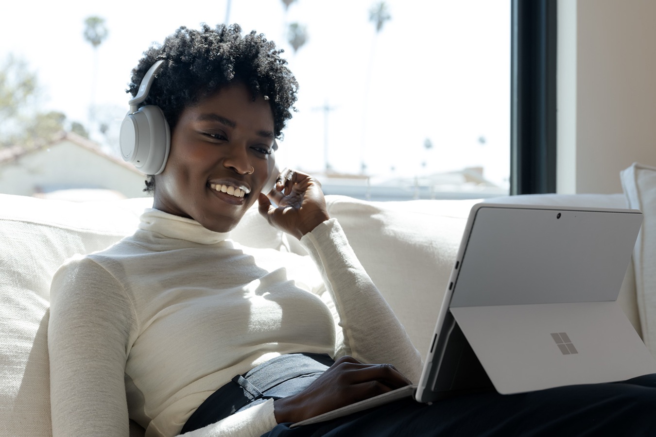 A woman, sitting on a sofa, uses her Surface Headphones 2 to interact with Surface Pro 8