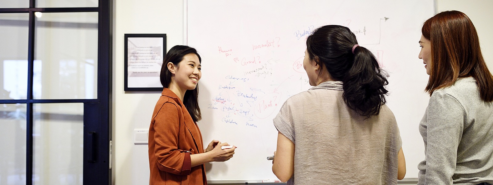 Women brainstorming in an office standing around a whiteboard.