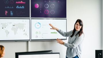 Una persona presentando datos en gráficos y tablas que se muestran en cuatro pantallas grandes en una pared detrás de ellos.