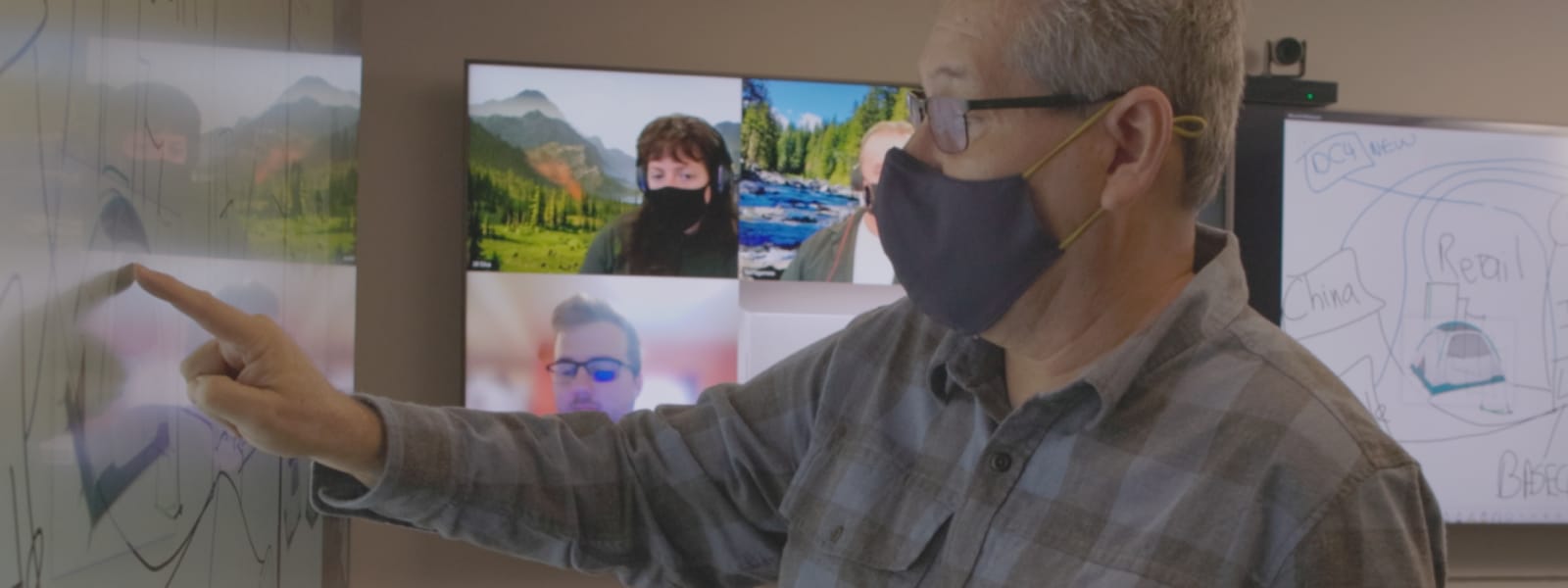 A person wearing a mask pointing to a whiteboard for a Teams video call
