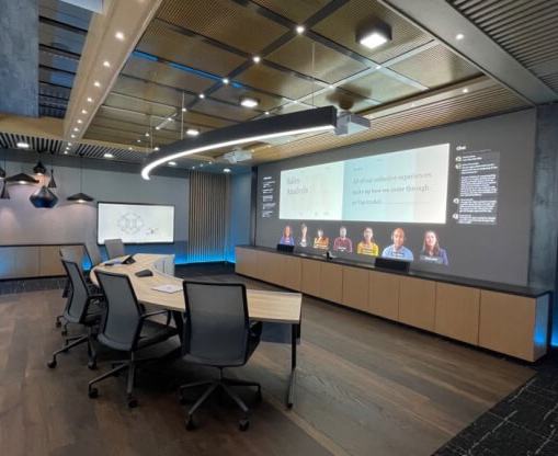 Empty conference room with half-circle table with chairs facing a large, wall-mounted screen showing a Microsoft Teams meeting.