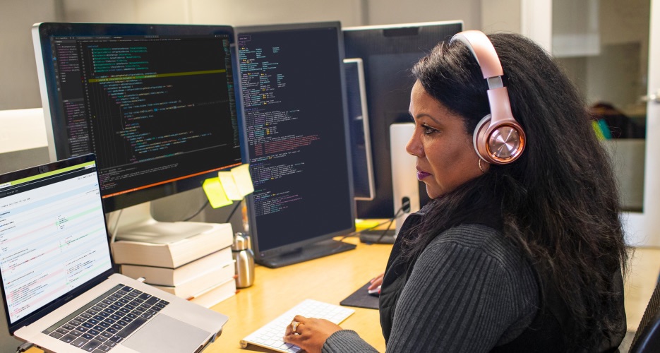 Henkilö käyttää kuulokkeet päässään kannettavaa tietokonetta, joka on yhdistetty useisiin näyttöihin.