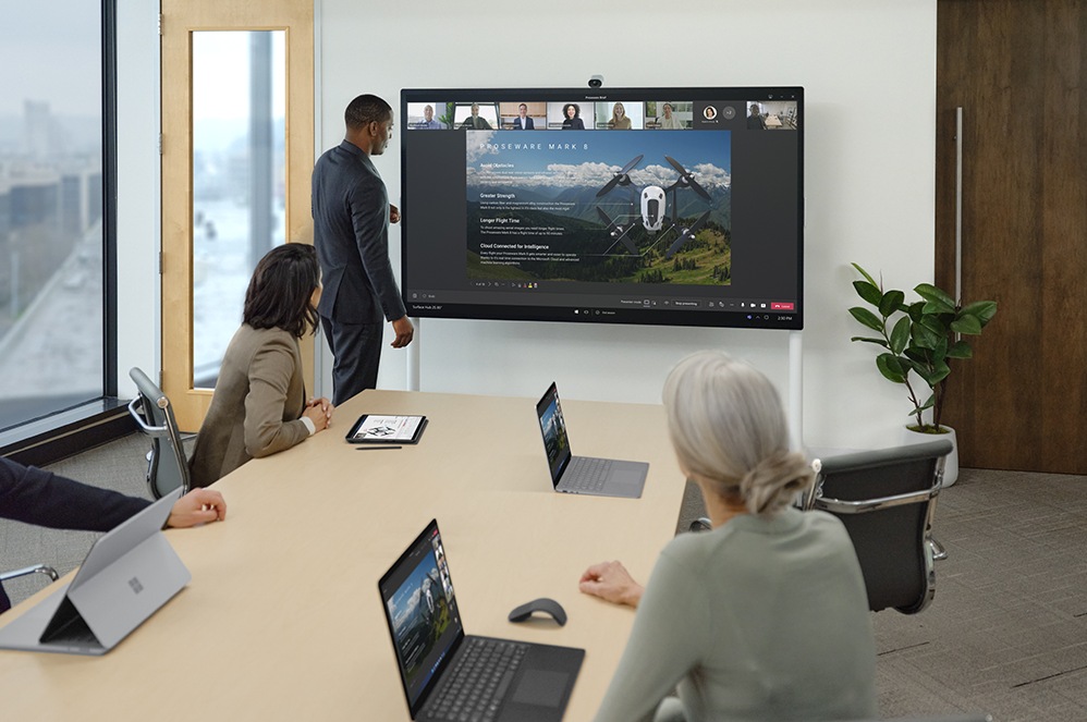 Medewerkers op kantoor en thuis bekijken samen een presentatie via Microsoft Teams met behulp van Surface Hub 2 Smart Camera en de Surface Hub 2S voor zakelijk gebruik.