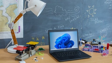 An open laptop sits on a table in a school classroom surrounded by science supplies with a blackboard in the background.