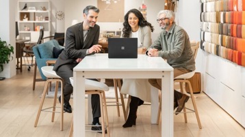 A group of people sit at a table talking with people in a Microsoft Teams meeting.